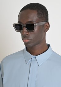 Graufarbene Sonnenbrille mit einem klobigen, klaren Acetatrahmen. Schwarze Gläser und rechteckige Form. Kombiniert mit einem hellblauen Hemd mit Kragen.