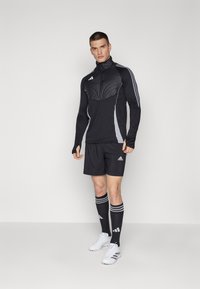Haut de sport noir à manches longues avec un panneau texturé sur la poitrine, shorts assortis, chaussettes noires montantes avec des rayures blanches et chaussures de sport blanches.