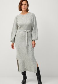 Abito lavorato a maglia grigio chiaro con maniche lunghe e voluminose, cintura legata in vita e spacco laterale. Tessuto morbido con motivo a coste. Stivali neri fino al ginocchio.