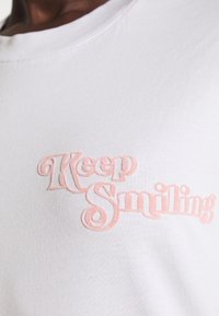 Chemise blanche avec le texte en relief rose "Keep Smiling" sur la poitrine, portée par une personne à la peau foncée.