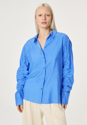 Vrouw draagt een losse, felblauwe blouse met knopen en gerimpelde mouwen, en crèmekleurige broek, staand tegen een effen lichte achtergrond.