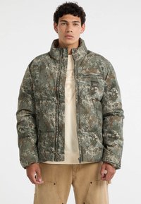 Giacca a vento in design camouflage con colori verde, marrone e crema. Presenta una chiusura a zip, colletto alto e una tasca sul petto.