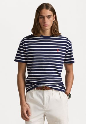 Camiseta de algodón de manga corta a rayas en azul marino y blanco. Presenta un pequeño logo rojo en el pecho. Combinada con pantalones cortos de color claro.