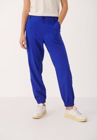 Blå, lätta, avslappnat sittande byxor med elastiska muddar och knäppning, i kombination med vita sneakers på en vit bakgrund.