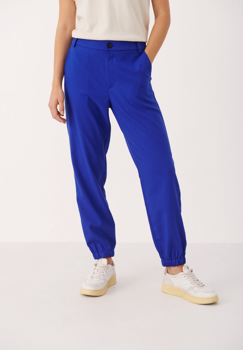 Blå, lätta, avslappnat sittande byxor med elastiska muddar och knäppning, i kombination med vita sneakers på en vit bakgrund.