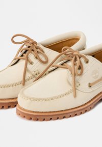 Μπεζ παπούτσια μοκασίνου από δέρμα με καφέ κορδόνια, επενδυμένους γιακάδες, ραμμένες λεπτομέρειες και ανθεκτικές καφέ ελαστικές σόλες.