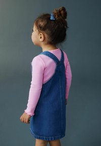 Vestito salopette di jeans con bretelle regolabili, abbinato a una maglietta rosa a maniche lunghe con polsini arricciati, visto da dietro.