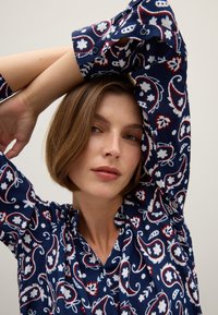 Marineblaues, gemustertes Hemd mit Paisleymuster in Weiß und Rot. Weicher Stoff, Knopfverschluss mit Kragen und hochgekrempelten Ärmeln.