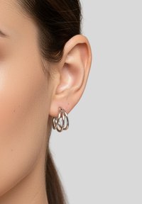Tre orecchini a cerchio intrecciati in argento con una finitura lucida, esposti su uno sfondo neutro vicino all'orecchio di un modello.
