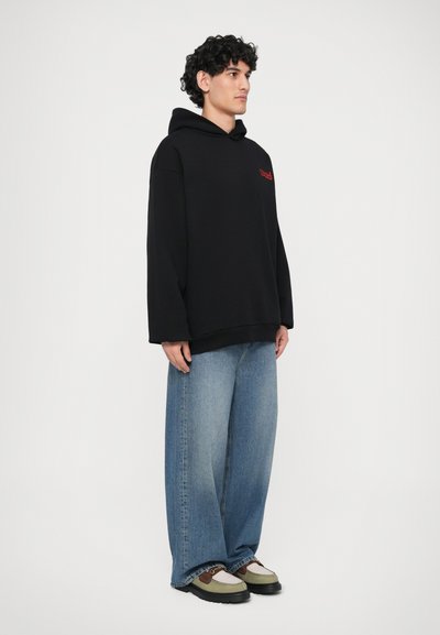 Schwarze oversized Kapuzenjacke mit einem roten Logo, kombiniert mit lockeren blauen Jeans. Die Schuhe zeigen eine Mischung aus grünen, beigen und schwarzen Farben.