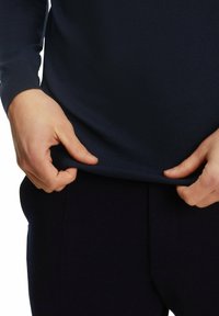 Marineblauer Strickpullover mit gerippten Bündchen und Saum, Hände, die den Saum greifen und die Textur und Passform des Materials gegen die dunklen Hosen betonen.