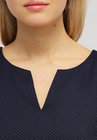 Námořnická modrá texturovaná blůza s výstřihem do V, zdobená malým kostkovaným vzorem a hladkým povrchem.