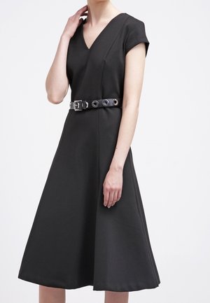 Femme portant une robe noire ligne A à col en V, manches courtes, avec une ceinture à œillets argentée sur fond blanc.