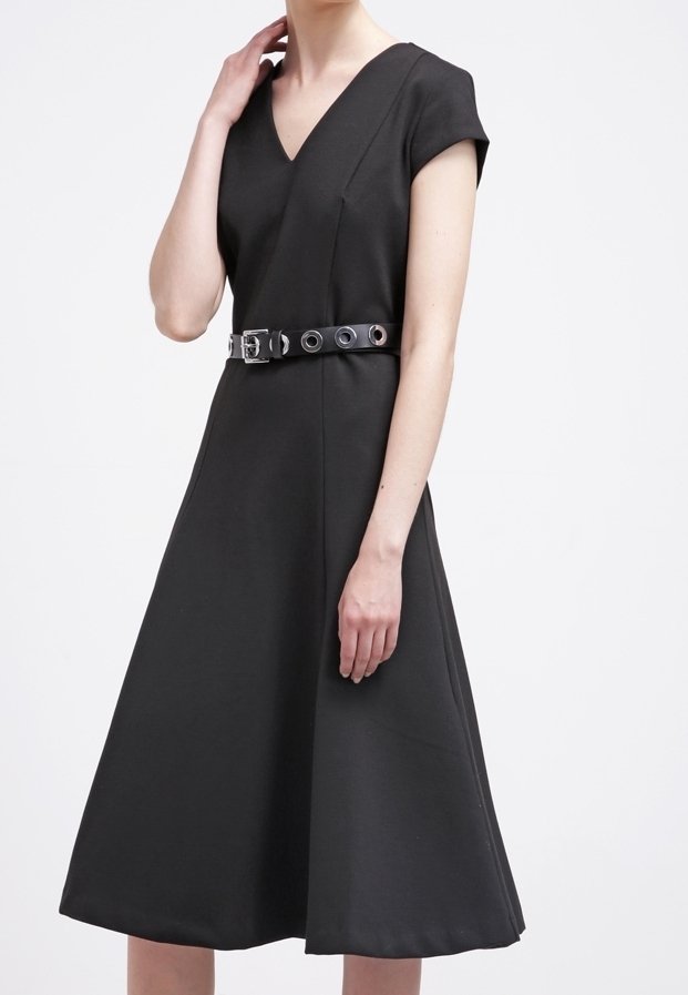 Femme portant une robe noire ligne A à col en V, manches courtes, avec une ceinture à œillets argentée sur fond blanc.