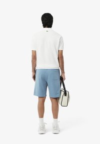 Camisa polo blanca de manga corta, pantalones cortos azul claro y lleva un bolso pequeño color crema con acentos en negro. Ropa deportiva casual.