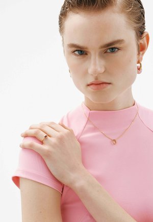 Collar de oro con un colgante circular, anillo de oro con una piedra preciosa, blusa ajustada rosa, piel suave y cabello mojado, centrado en los rasgos faciales.