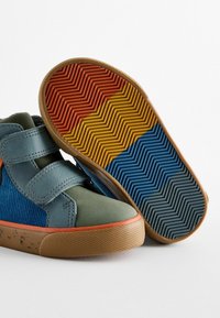 Scarpe alte in tessuto blu e verde, con suola in gomma beige e accenti arancioni. La suola presenta un motivo testurizzato in rosso, giallo e blu.