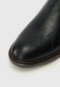 Chaussure en cuir noir avec un bout pointu, texture lisse et détails en cuir marron cousus le long de la semelle. Motif grainé subtil sur le cuir.