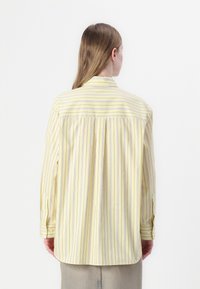 Camicia gialla a righe con bottoni, maniche lunghe e dettaglio plissettato sul retro. Presenta righe a contrasto viola e blu. Tessuto morbido.