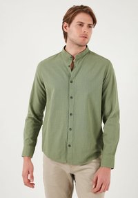 Chemise verte à manches longues avec un col montant, dotée d'un tissu texturé et de boutons noirs sur le devant. Design lisse et ajusté.