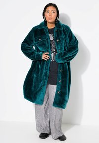 Cappotto oversized in velluto teal con due tasche sul petto e chiusura a bottoni, abbinato a pantaloni larghi grigi con paillettes e scarpe nere.