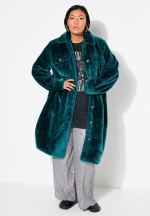 Abrigo de peluche en tono verde azulado, de corte oversized, con dos bolsillos en el pecho y cierre de botones, combinado con pantalones de pierna ancha en gris con lentejuelas y zapatos negros.