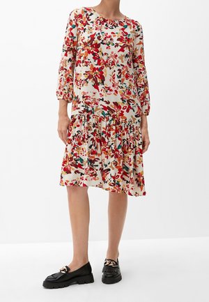 Vrouw die een losse bloemenjurk draagt met rode, oranje en groene bladeren, driekwart mouwen en zwarte loafers met gouden kettinkjes.