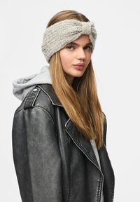 Jeune femme aux longs cheveux raides portant un bandeau gris en tricot, un sweat à capuche gris et une veste en cuir noire, regardant légèrement sur le côté.