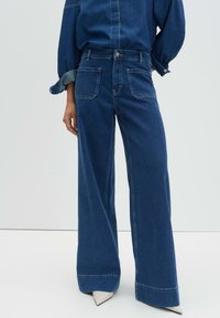 Femme portant un jean taille haute, large et bleu foncé, associé à une chemise en denim assortie et des chaussures blanches à bouts pointus.