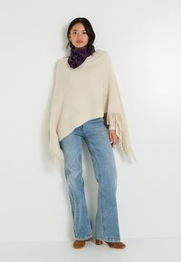 Poncho beige con bordes de flecos, combinado con jeans de campana azules claros y una bufanda morada de estampado. Material de punto texturizado, corte relajado.