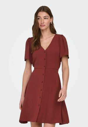 Kvinde iført en rustrød, kortærmet kjole med knapper, V-hals og diskrete balloneærmer, stående foran en hvid baggrund.