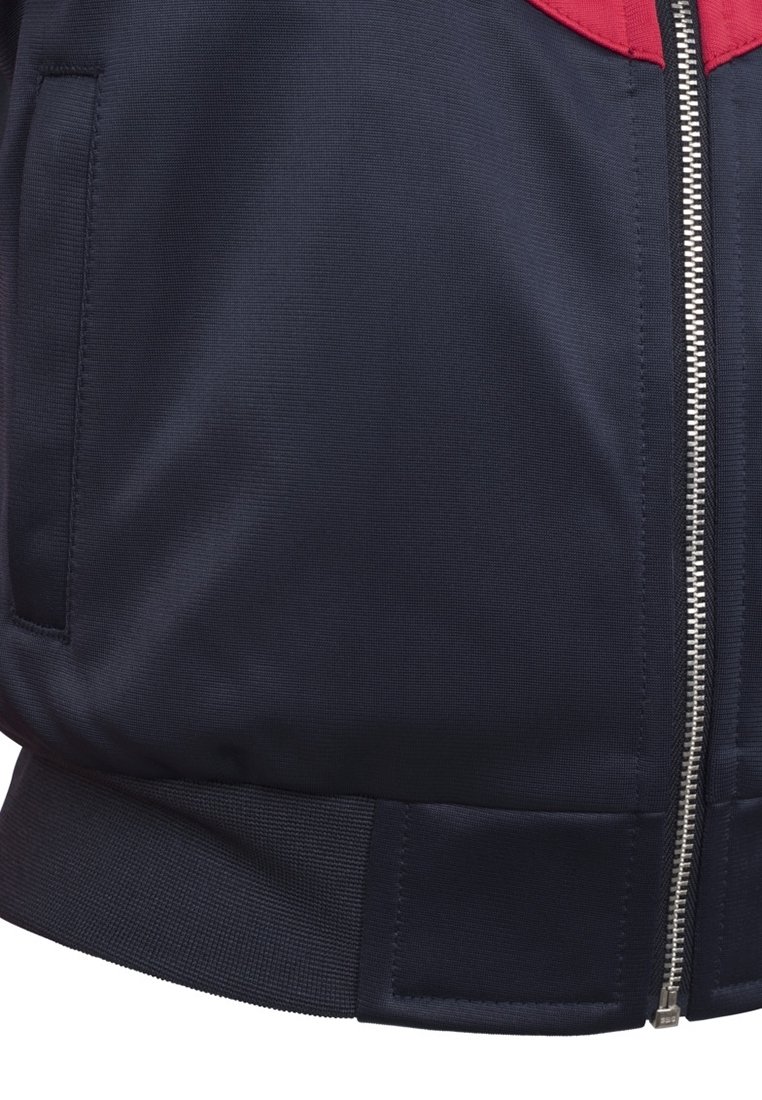 Close-up van de onderkant van een donkerblauwe jas met een zilveren rits, zijzak en een rode stoffen detail aan de rand van de kraag.