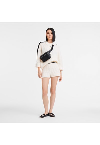 Cremfarbener Kabelstrick-Pullover mit schwarzen Streifen und Knopfdetails, kombiniert mit cremfarbenen Shorts. Schwarze Umhängetasche und flache schwarze Schuhe.