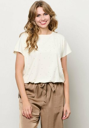 Lachende vrouw met golvend blond haar draagt een wit topje met korte mouwen en parels, gecombineerd met losse satijnen beige broek met een gestrikte taille.