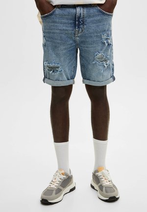 Short en denim avec revers roulés et patchs usés, porté avec des chaussettes blanches et des baskets grises sur une personne debout.