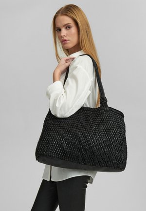 Pikkade blondide juustega naine, kes kannab valget särki ja musti pükse, hoides õlal suurt musta punutud käekotti.