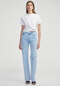 Witte t-shirt met korte mouwen en een geknoopt detail bij de taille, gecombineerd met lichtblauwe rechte jeans met vervaagde strepen en versleten accenten.