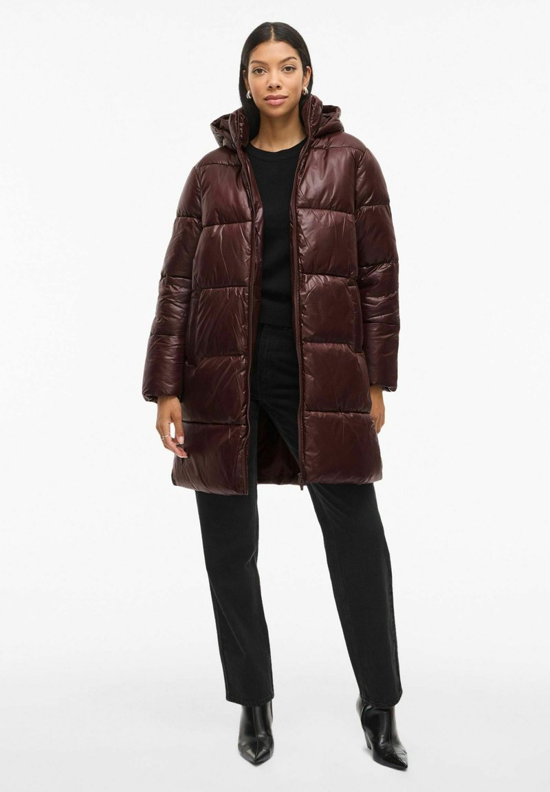 Cappotto imbottito bordeaux con cappuccio, caratterizzato da una chiusura frontale con zip, ampie sezioni trapuntate e una finitura lucida, abbinato a pantaloni neri e stivali con tacco.