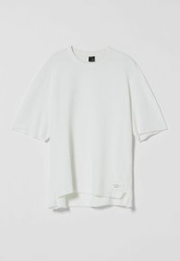 Bílé oversized tričko s krátkým rukávem, kulatým výstřihem, s nárazy na surovém lemu a malou nálepkou našitou v levém dolním rohu.