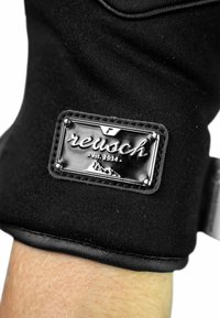 Black fabric glove featuring a glossy label with the brand name "reusch" and establishment year, set against a textured wrist area.