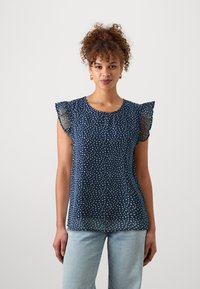 Mornarska bluza z belimi pikami, prosojnimi ruffled rokavi, okroglo ovratnikom in lahkim materialom; v kombinaciji s svetlo modrimi trenirkami.