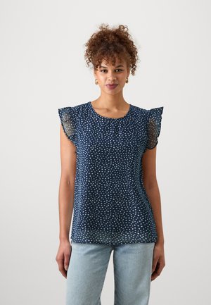 Navy blouse with white speckled pattern, sheer ruffled sleeves, round neckline, and lightweight fabric; paired with light blue jeans.