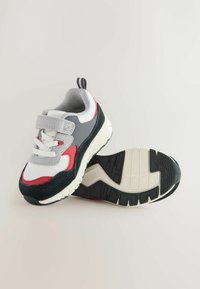 Sportssko med en hvid, grå og rød farveskema. Har syntetisk overdel, velcro-strop og en struktureret sort gummisål.