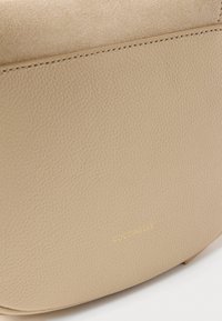 Beige lederen tas met een gestructureerd oppervlak, suède bovenklep en fijne stiksels, met een klein goudkleurig "COCCINELLE" logo in reliëf.