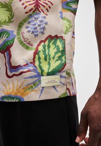 Kortærmet, mønstret skjorte i beige med grønne, røde og blå blomsterdesigns. Har en logotag i kanten, der viser branddetaljer.