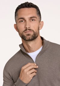 Brauner Reißverschluss-Pullover aus Rippmaterial, mit Stehkragen und einem weißen Shirt darunter. Texturierte Oberfläche mit subtilen Designdetails.