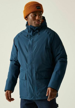 Mężczyzna w niebieskiej zimowej kurtce z kapturem i pomarańczowej dzianinowej czapce, stoi z rozluźnionymi rękami, patrząc w bok na jednolitym tle.