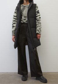 Svart pufferväst över en mönstrad tröja, kombinerad med mörka vida byxor och kraftiga svarta stövlar. Släta texturer med varierade mönster.