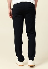 Hombre con pantalones rectos negros, zapatillas blancas y una camisa blanca de pie contra un fondo crema, visto desde atrás.