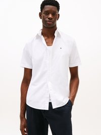Un giovane uomo veste una camicia bianca a maniche corte sopra una canotta bianca, con pantaloni scuri e una mano in tasca.